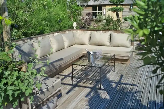 Salon de jardin avec banquette et table basse sur une terrasse en bois entourée de végétation